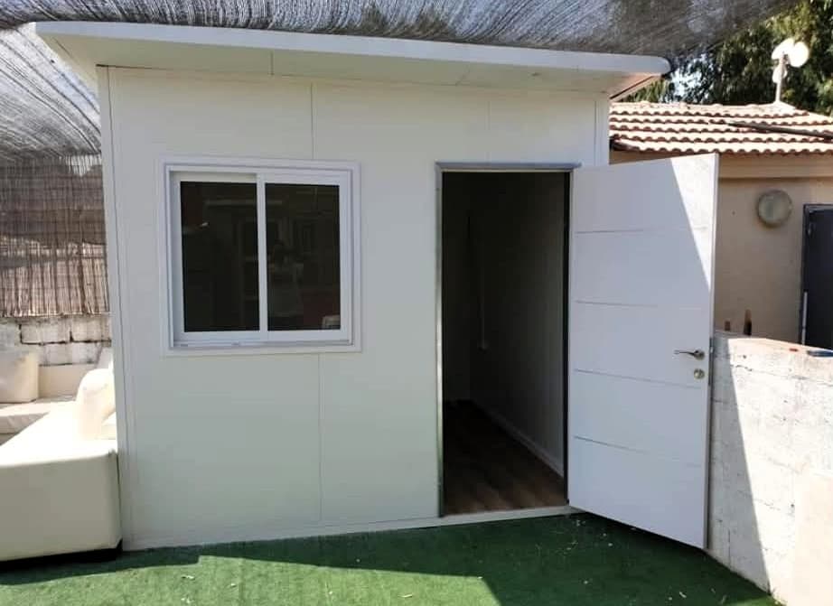 מבנה יביל לבן קטן עם דלת פתוחה וחלון זכוכית, ניצב על דשא סינטטי ירוק.